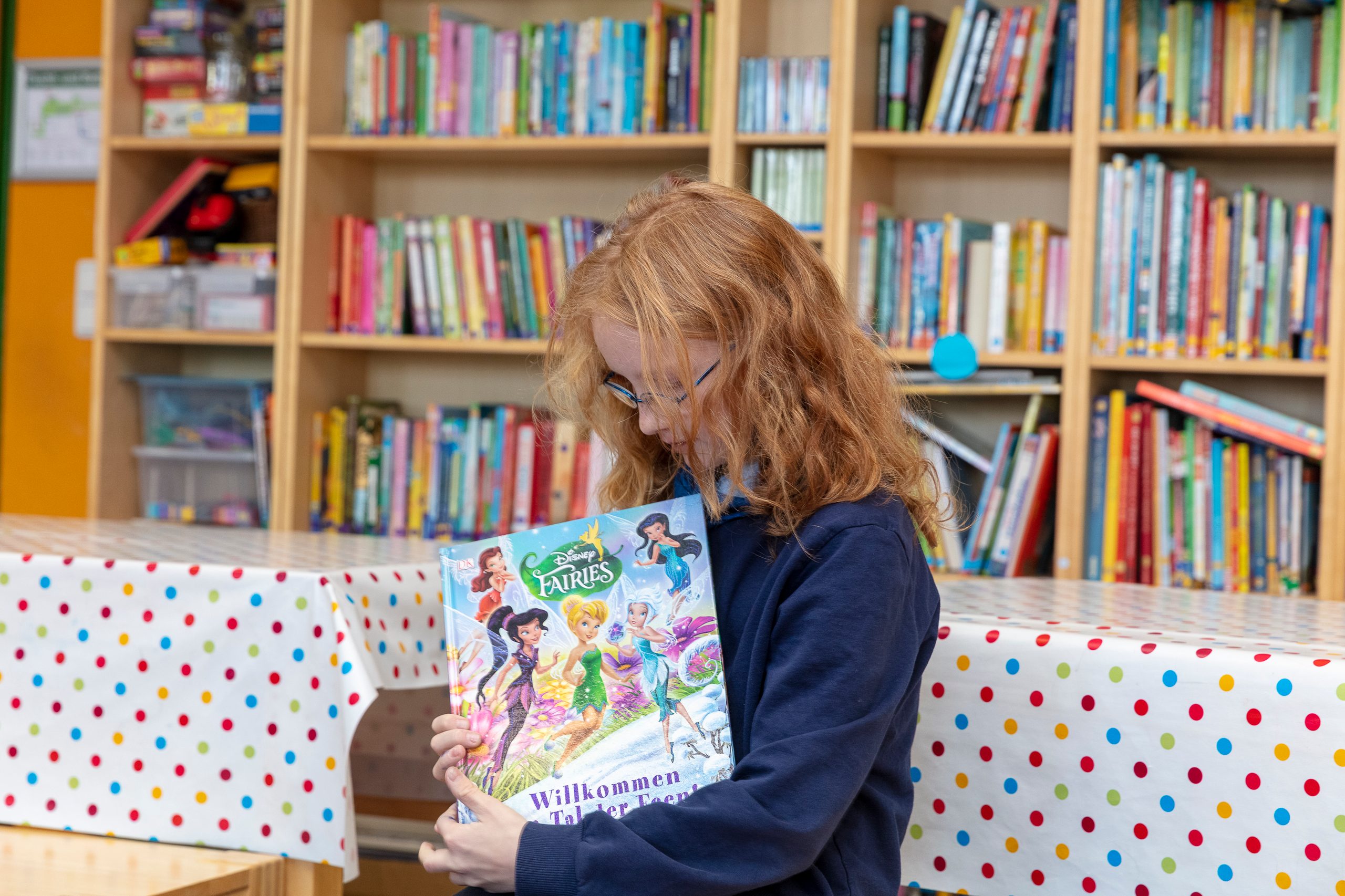 Anne sitzt vor dem Tisch, der vor dem Bücherregal steht. Sie hält das Buchcover sichtbar und schaut es selbst an.