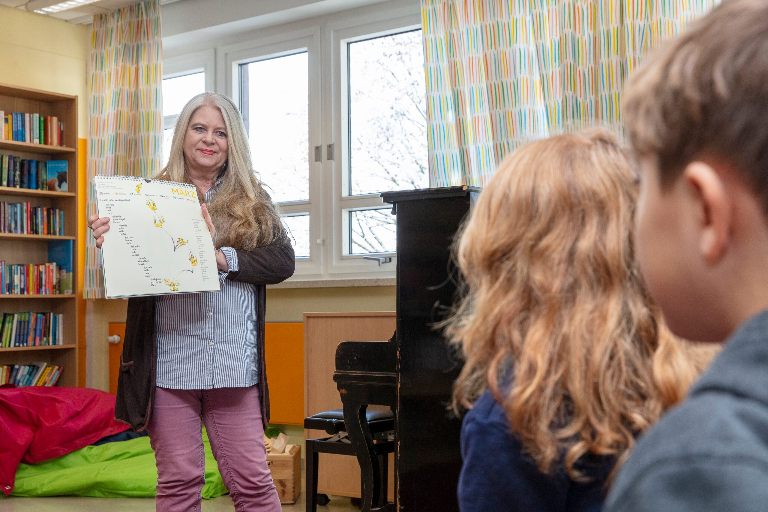 Frau Pelz steht neben einem schwarzen Klavier und zeigt den Kindern ein Kalenderblatt.