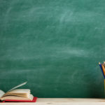 Klassenzimmer mit Tafel und im Vordergrund Buch und Stifte