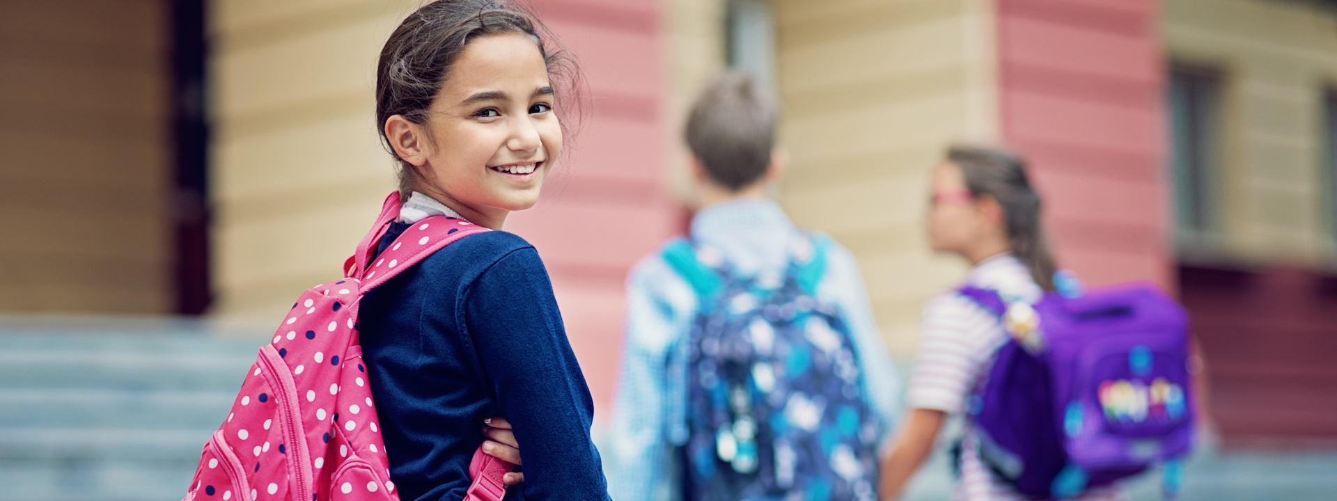 Schulmädchen mit Rucksack steht lächelnd vor Schule