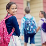 Schulmädchen mit Rucksack steht lächelnd vor Schule